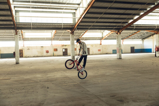 Male BMX rider balancing on rear wheel beneath exposed trusses inside warehouse on red BMX bicycle - Powered by Adobe