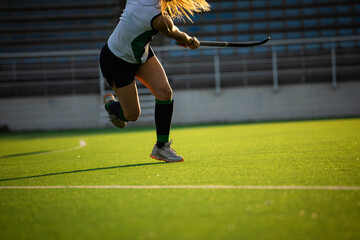 Teenage girl field hockey player sprinting across artificial turf pitch and swinging hockey stick