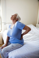 Senior woman is sitting on bed by vertical blinds and curtains, pressing lower back and stretching