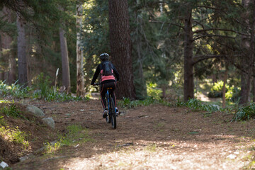 Naklejka premium Female cyclist riding narrow dirt trail in dense forest on mountain bike with helmet, backpack