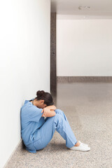 Asian woman in blue scrubs sitting on floor of hospital corridor leaning against wall, copy space