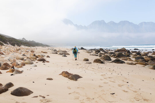 Woman walking on sandy beach wearing wide-brim hat and carrying backpack using trekking poles