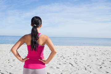 Naklejka premium Asian woman standing on white sand beach facing calm blue sea wearing pink top and leggings