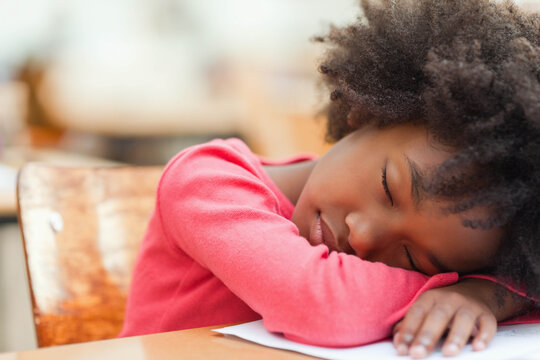 African American girl student resting head on folded arms on wooden desk with paper in classroom