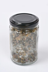 A glass jar with a black lid is filled with capers in salt on a white background