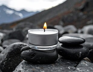 A single white candle flame illuminates a serene scene of stacked smooth black stones against a blurred mountain backdrop