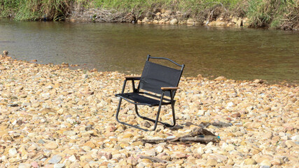 Fototapeta premium Black folding camping chair placed on rocky riverbed with clear water, perfect for outdoor adventure, travel, relaxation, nature, and summer vacation background