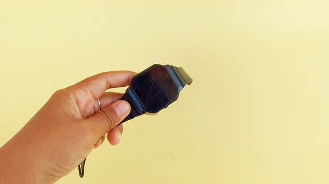 a woman's hand holding a black watch on a yellow background - Powered by Adobe