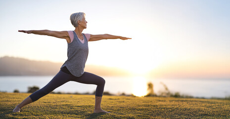 Mature woman practicing warrior ii pose on grass, silhouetted against sunset ocean, embodying wellness and inner strength with copy space