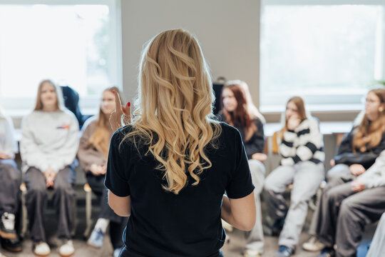A blonde teacher with curly hair addresses a group of attentive students seated in a bright classroom, fostering discussion, engagement, and interactive learning - Powered by Adobe