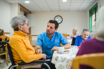 Young male caregiver providing assistance and talking with an elderly woman in a wheelchair during an activity at a retirement home