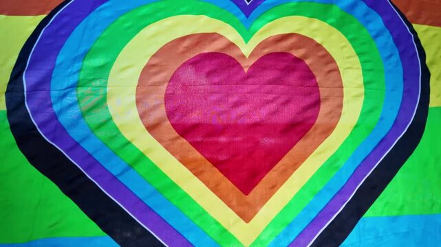 Colorful rainbow flag with a heart shape waving in the wind in slow motion, a symbol of love and freedom for the LGBTQ+ community during pride month celebrations and gay pride parades
