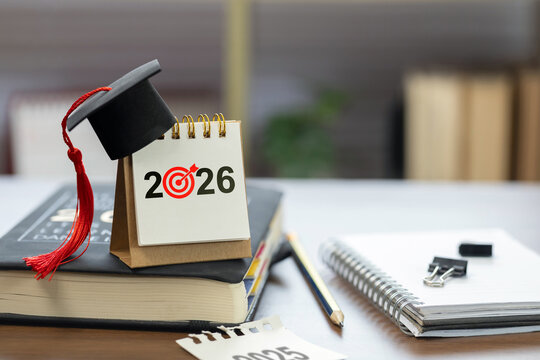 Desk calendar marked 2026 with a red target icon and graduation cap on books beside notebook and pencil, symbolizing academic planning, study roadmap, college goals, graduation success in the new year