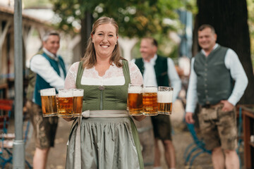 Traditional waitress in dirndl serves mugs of beer to cheerful friends in Bavarian costumes celebrating Oktoberfest in an outdoor beer garden