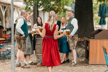 Smiling waitress in Bavarian dirndl serving mugs of beer to friends at Oktoberfest enjoying leisure friendship and festive beer garden culture