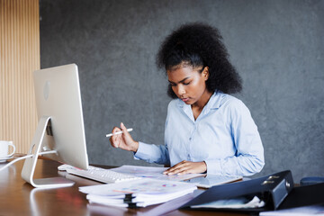 A young professional woman works at her desk with a computer, reviewing charts and opening a file folder, reflecting focus, organization, productivity, and dedication in a modern business environment.