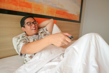 Happy young man with a remote controller is lying in bed and changing channels