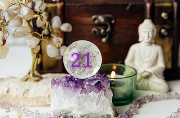 Number twenty-one gemstone sphere or crystal ball known as crystallum orbis. Natural clear quartz ball on stand on crystal geode home, candle burning. Predictions concept.
