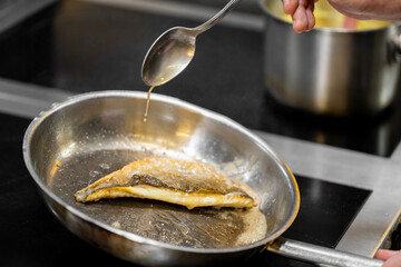 Chef drizzling sauce over pan-seared fish fillet in stainless steel skillet. Golden crispy skin, professional kitchen setting, gourmet cooking in progress.
