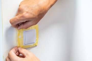 Man covering a light switch with masking tape before painting the wall white. Home renovation, surface protection and careful preparation during interior work.