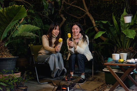 Happy women friends enjoy winter outdoor camp forest night, people gather warm fire roasting corn in cozy sweaters, creating cheerful winter memories