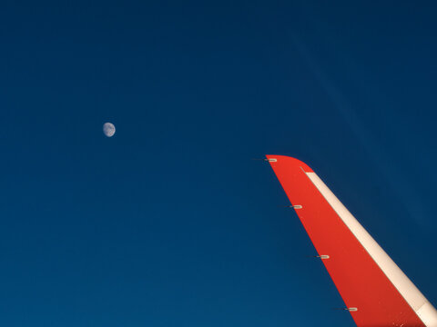 An aerial view from an airplane window, capturing the winglet against a deep blue sky with the moon visible in the distance.