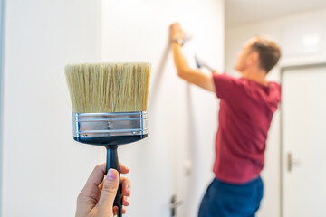 Close-up of a hand holding a paintbrush with a man painting the wall in the background. Concept of manual work, home renovation and attention to detail during interior improvement.