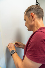 Man covering a light switch with masking tape before painting the wall white. Home renovation, surface protection and careful preparation during interior work.