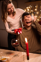 young couple in love celebrating valentine's day at home, have romantic dinner. woman covering man eyes and giving gift box on birthday in home.