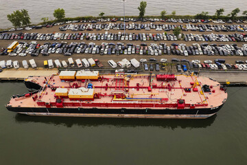 Brooklyn, United States - 29 August 2021: Aerial view of a vibrant tanker ship docked beside a vast parking lot filled with hundreds of cars.