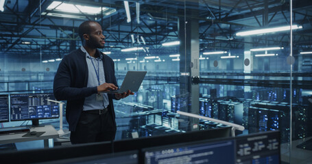 Black Male Software Developer Analyze System Performance Metrics on a Laptop Inside a Modern Data Center. Young Manager Ensuring Efficient Operations for Critical Infrastructure