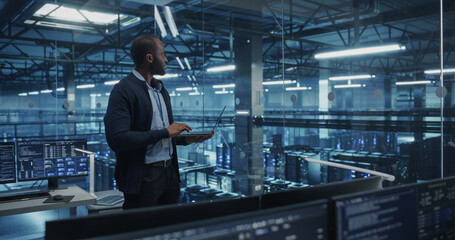Black Male Software Developer Analyze System Performance Metrics on a Laptop Inside a Modern Data Center. Young Manager Ensuring Efficient Operations for Critical Infrastructure