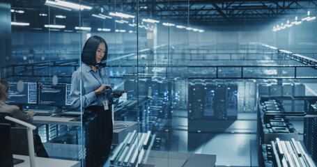 Asian Female Data Analyst Studies Traffic Logs on Her Laptop Computer, Standing in a Modern Data Center with Server Systems. Young Woman Searching for Patterns to Improve Online Data Flow