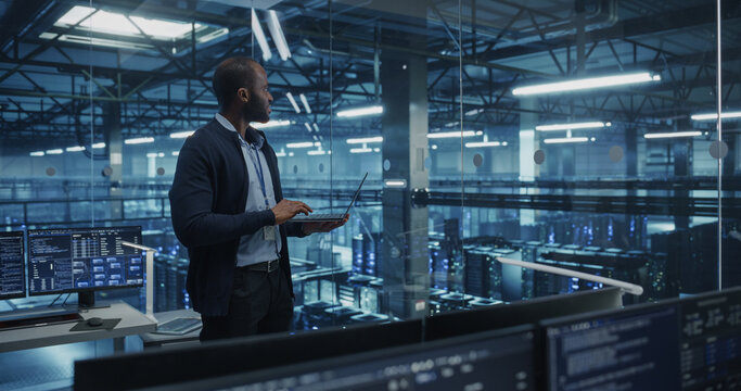 Black Male Software Developer Analyze System Performance Metrics on a Laptop Inside a Modern Data Center. Young Manager Ensuring Efficient Operations for Critical Infrastructure