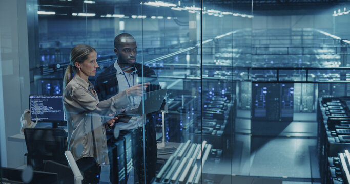 Male DevOps Engineer and Female IT Consultant Work on a Laptop Computer, Standing in an Office Above the Rows of Servers in a Data Center, Developing Automation Scripts for Artificial Intelligence
