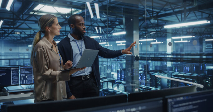 Male Cloud Architect and Female Database Engineer Review Data Storage Protocols on a Shared Laptop in a Data Center. Team Strategizing Ways to Improve Service for Enterprise Clients