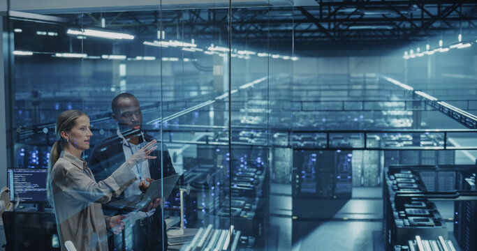 Male DevOps Engineer and Female IT Consultant Work on a Laptop Computer, Standing in an Office Above the Rows of Servers in a Data Center, Developing Automation Scripts for Artificial Intelligence - Powered by Adobe