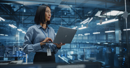 Positive Female Infrastructure Architect Reviewing Virtualization Strategies on a Laptop Computer, Standing in Office Next to Towering Racks of Servers, Planning to Scale Client Cloud Solutions