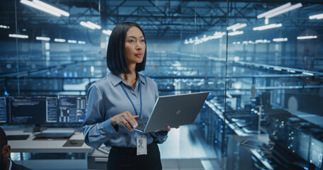 Asian Software Developer Analyze System Performance Metrics on a Laptop Inside a Modern Data Center. Young Female Manager Ensuring Efficient Operations for Critical Infrastructure