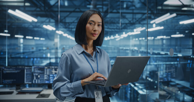 Portrait of a Data Center Administrator Standing in a High-Tech Server Room, Optimizing Server Configurations on a Laptop Computer. Female Specialists Looking at Camera and Smiles - Powered by Adobe