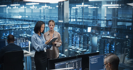 Two Diverse Female Data Center Engineers, an Asian and Caucasian Women, Collaborate Using a Tablet Computer in a Glass Office Next to a Futuristic Server Room, Analyzing Software Data on Screen