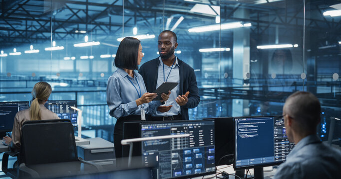 Male DevOps Engineer and Female IT Consultant Using a Tablet Computer, Standing in an Office Above the Rows of Servers in a Data Center, Developing Automation Scripts for Artificial Intelligence - Powered by Adobe