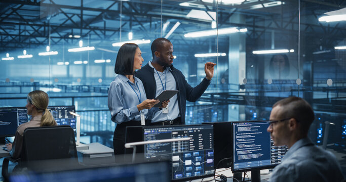 Male DevOps Engineer and Female IT Consultant Using a Tablet Computer, Standing in an Office Above the Rows of Servers in a Data Center, Developing Automation Scripts for Artificial Intelligence