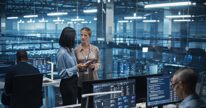 In a Modern Data Center, Caucasian Female Data Scientist and Asian Female AI Specialist Analyze Algorithms on a Tablet Computer. Specialists Exploring Optimizations for Processing Large Datasets