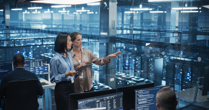 In a Modern Data Center, Caucasian Female Data Scientist and Asian Female AI Specialist Analyze Algorithms on a Tablet Computer. Specialists Exploring Optimizations for Processing Large Datasets