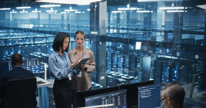 Two Diverse Female Data Center Engineers, an Asian and Caucasian Women, Collaborate Using a Tablet Computer in a Glass Office Next to a Futuristic Server Room, Analyzing Software Data on Screen