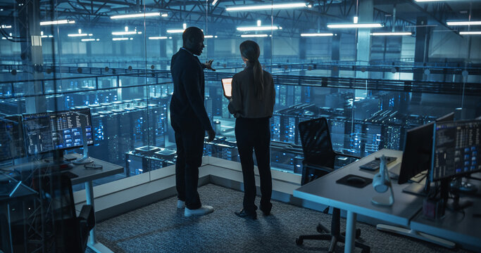 Male DevOps Engineer and Female IT Consultant Work on a Tablet Computer, Standing in Office Above the Rows of Servers in a Data Center, Working on Research and Development of Artificial Intelligence