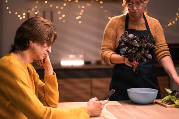 family evening scene in kitchen husband working overtime sitting at table while wife prepares fresh salad for dinner together home. life work balance