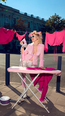 Woman in pink outfit with curlers joyfully ironing clothes on street with playful energy. Concept of surreal editorial, lifestyle branding, advertising creativity, modern fashion art.
