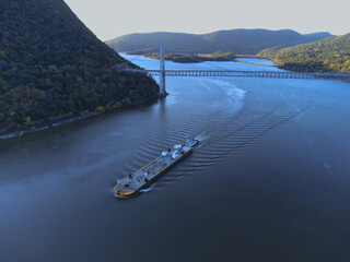 Fort Montgomery, United States - 17 October 2021: Aerial view of a long barge gliding smoothly on the Hudson River, under the Bear Mountain Bridge, against the backdrop of forested hills.